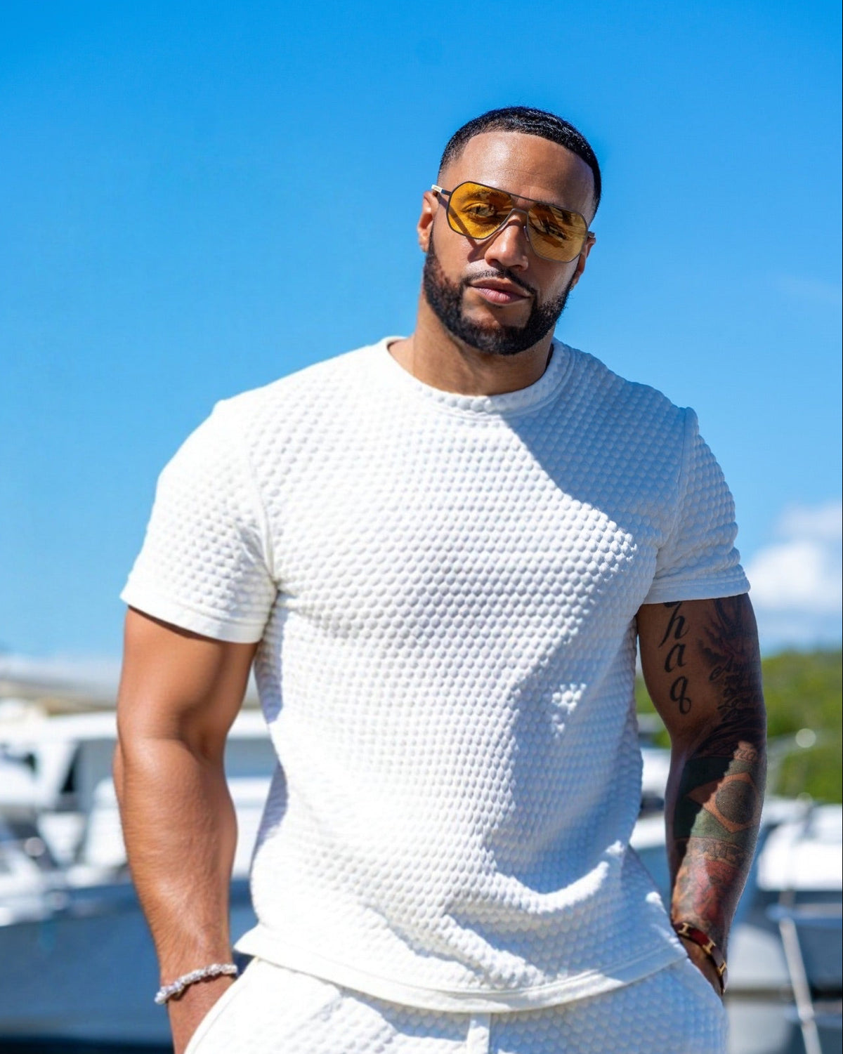 Man wearing a white textured t-shirt and pants with sunglasses, standing by a marina.