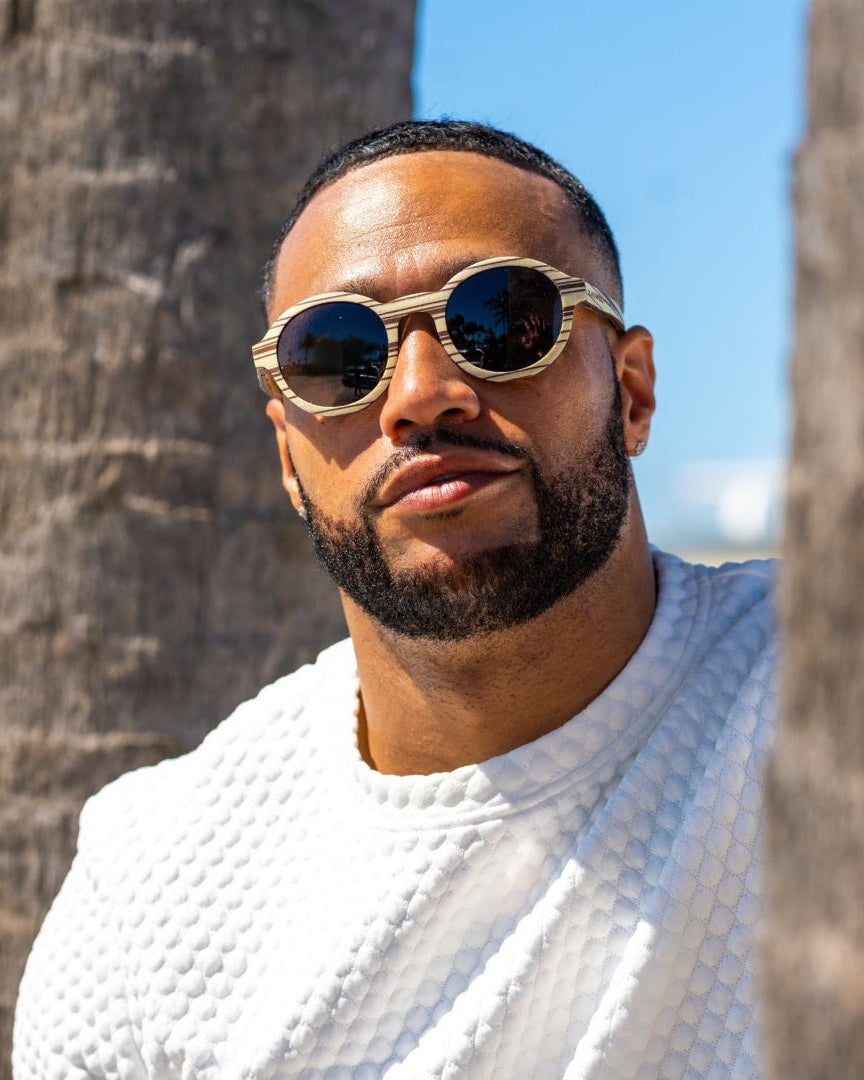 Man wearing white zebrawood sunglasses and a white textured shirt standing between two palm trees.