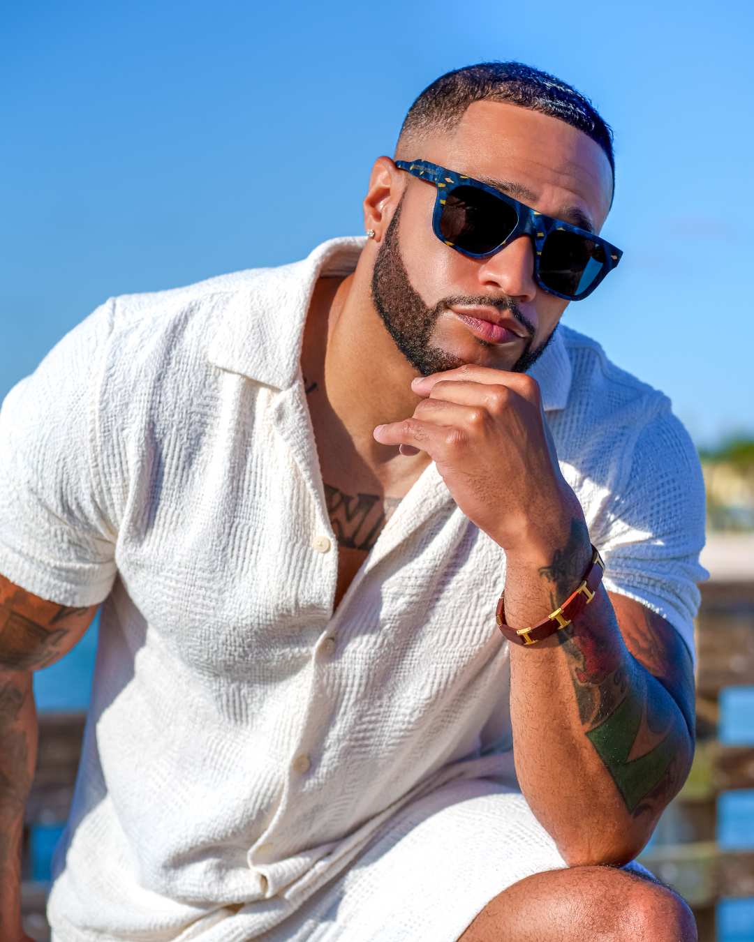 Man wearing stylish wood sunglasses  by Blackwood Premium and a white shirt with tattoos, sitting outdoors.